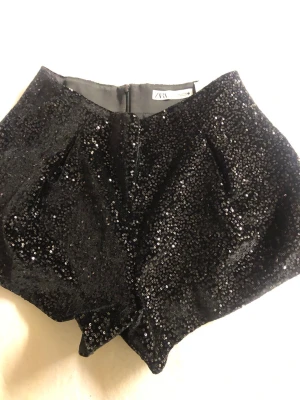 Svarta paljettshorts från Zara - Glittriga svarta shorts från Zara med paljetter över hela ytan. Modellen är högmidjad och har en snygg, lite lös passform. Perfekta för att sticka ut och skapa en cool look.