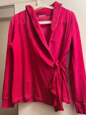 Rosa omlottkofta i velour - Säljer en rosa omlottkofta i mjuk velour med knytband i midjan och långa ärmar. Koftan har en avslappnad passform och bred krage, perfekt för lager-på-lager och en trendig look. Se bilder för defekter/ små hål (2 st).