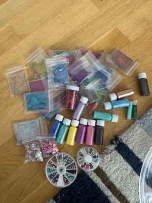 Stort kit med nageldekorationer & glitter - Säljer ett stort kit med nageldekorationer, glitter, paljetter och pigment i många olika färger. Innehåller små burkar och påsar med glitter i blått, rosa, lila, grönt, guld, silver och mer, samt dekorationshjul med stenar och figurer. Perfekt för kreativa nageldesigner, smink eller ljustillverkning