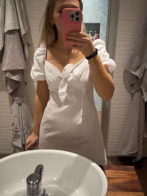 Vit kort studentklänning  - Söt vit kortklänning med puffärmar och knyt framtill. Klänningen har en v-ringad urringning och är tillverkad i ett lätt bomullstyg. Perfekt för varma dagar och har en feminin, trendig look. perfekt till studenten/ midsommar. har aldrig kommit till användning och köpte den här på plick!  