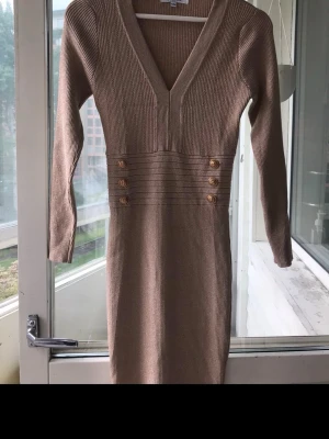 Beige ribbad klänning med knappar - Stilren beige ribbad klänning med v-ringning och dekorativa guldknappar i midjan. Klänningen har lång ärm och figurnära passform, perfekt för dig som gillar en klassisk look med snygga detaljer.