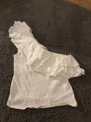 Vit oneshoulder topp med volang - Säljer en vit oneshoulder topp med bred volangdetalj över axeln. Toppen har en figurnära passform och är tillverkad i ett mjukt, lätt material som känns skönt mot huden. Perfekt för dig som vill sticka ut med en unik och trendig look.