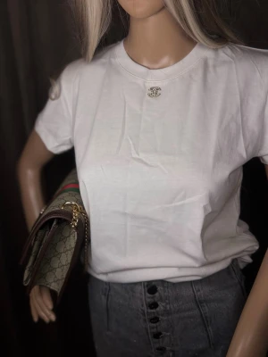 Vit basic t-shirt med pärlbroderad detalj - En klassisk vit t-shirt i bomull med rund hals och korta ärmar. På bröstet sitter en pärlbroderad detalj som ger en exklusiv touch. Perfekt att styla med jeans eller kjol för en clean och trendig look.