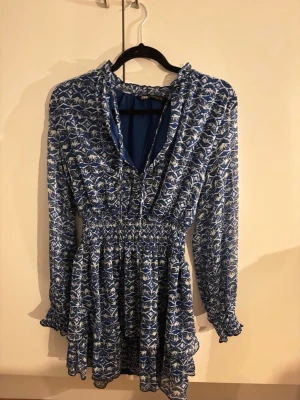 Blåvit mönstrad kortklänning Zara - Kortklänning från Zara i blått och vitt med grafiskt mönster. Klänningen har långärmade ärmar med smock vid ärmslut, markerad midja och lätt volang i kjolen. Tillverkad i ett tunt, flowy material som ger en luftig känsla.