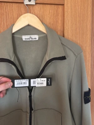 Beige ziptröja från Stone Island - Stilren olivgrön ziptröja från Stone Island med svart dragkedja och klassisk logotyp-patch på ärmen. Tröjan har två stora fickor framtill och ribbade muddar. Perfekt för en avslappnad och trendig look.