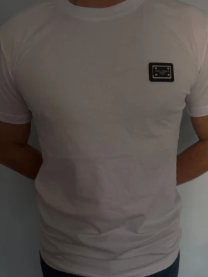 Vit Dolce & Gabbana t-shirt - Snygg vit t-shirt från Dolce & Gabbana med klassisk passform och korta ärmar. T-shirten har en svart logopatch på bröstet och är tillverkad i mjuk bomull. Perfekt för en clean och stilren look.