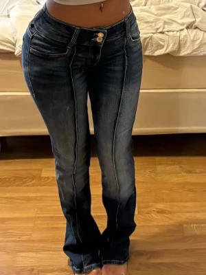 Mörkblå bootcut jeans med dubbla knappar - Snygga mörkblå bootcut jeans med markerade sömmar och dubbla knappar i midjan. Jeansen har klassiska fem fickor och lätt slitna detaljer längs benen. Perfekta för en trendig och avslappnad look. Har ett litet hål på sidan annars inget fel