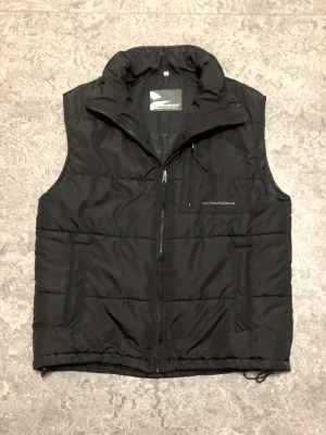 Svart Väst från Oatmandcoo - Stilren svart dunväst från Oatmandcoo med hög krage och dragkedja framtill. Västen har två sidofickor och en bröstficka med dragkedja. Perfekt för lager-på-lager och enkel att matcha med olika outfits.