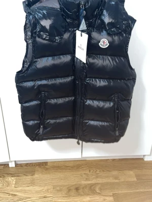 Svart dunväst från Moncler - Snygg svart dunväst från Moncler med glansig finish, huva och två dragkedjeförsedda fickor framtill. Västen har Moncler-logga på bröstet och quiltad design som ger en modern look. Perfekt för lager-på-lager-stil under kyliga dagar.