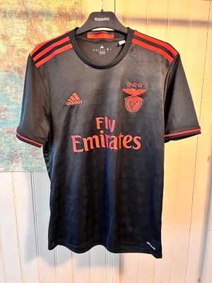 Benfica Adidas 2016/17 away shirt, size M - Good condition.  #lindelof #ederson #guedes #benfica #portugal #adidas #jimenez #grimaldo  Svart Benfica fotbollströja med röda detaljer. Tröjan har klubbens emblem och sponsorlogotyper tryckta på framsidan, samt ”Sagres” tryckt på baksidan.