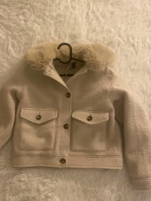 Beige cardiganjacka med fuskpäls - Snygg beige cardiganjacka med fluffig fuskpäls på kragen och stora fickor framtill med knappar. Jackan har en boxig passform och stängs med knappar. Perfekt för kyliga dagar och ger en trendig look.
