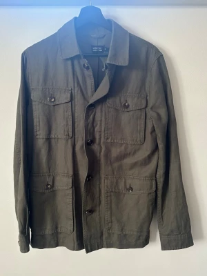 Olivgrön overshirt  - Stilren olivgrön overshirt från ett franskt märke med fyra stora fickor framtill och knäppning med svarta knappar. Rak passform och klassisk krage. Overshirten är helt ny och har endast provats. 