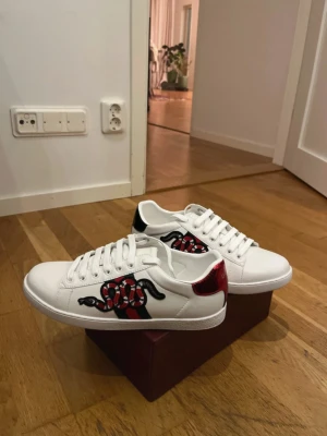 Gucci Ace vita sneakers med orm - Säljer ett par vita Gucci Ace sneakers i läder med ikonisk röd och svart orm-brodyr på sidan. Skorna har röda och svarta detaljer på hälen och klassisk låg profil. Snygga och trendiga sneakers med tydlig logga på sulan. Väldigt grish och pris går alltid att diskutera