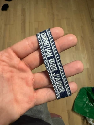 Blått Christian Dior J'Adior armband - Snyggt blått tygarmband från Christian Dior med texten 'Christian Dior J'Adior' i vitt. Armbandet har justerbar knytning med tofsar i ändarna och vita detaljer längs kanterna. Perfekt accessoar för att lyfta din outfit.