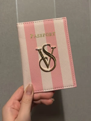 Victoria's Secret passfodral - Säljer ett snyggt passfodral från Victoria's Secret i rosa och ljusrosa ränder. Fodralet har ett stort guldfärgat VS-emblem på framsidan och är gjort i konstläder. Insidan har fack för boardingkort och pass, med text i guld. Perfekt för resan och supercute accessoar!