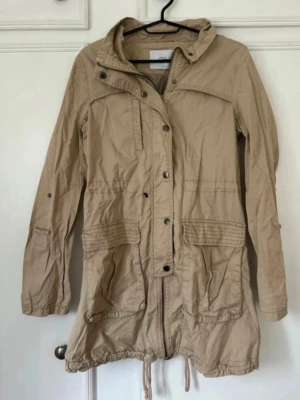 Beige parkasjacka med fickor - Stilren beige parkasjacka med dragsko i midjan och nederkant. Jackan har stora framfickor, knapp- och dragkedjestängning samt en hög krage. Perfekt för lager på lager och enkel att matcha med olika outfits.