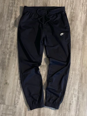 Mörkblåa Nike mjukisbyxor  - Säljer ett par mörkblå mjukisbyxor från Nike med klassisk vit broderad logga på vänster ben. Byxorna har dragsko i midjan, resår vid bensluten och sidofickor. Perfekta för chill dagar eller träning. Materialet känns mjukt och bekvämt.