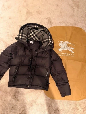 Burberry Svart vinter dunjacka  - Svart dunjacka från Burberry med klassiskt rutigt foder i huvan och dragkedja samt knappar framtill. Jackan har två fickor med dragkedja och en patch på ärmen. Perfekt för kalla dagar.