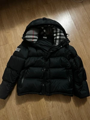 Svart pufferjacka från Burberry - Snygg svart pufferjacka från Burberry med klassiskt rutigt foder i huvan och dragkedja framtill. Jackan har två stora fickor, vadderad design och en patch med Burberry-logga på ärmen. Perfekt för kalla vinterdagar. Går att ta av armarna. 1:1