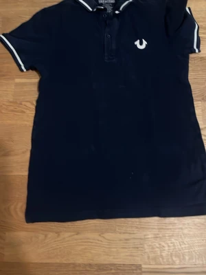 Marinblå piké från True Religion - Snygg marinblå pikétröja från True Religion med vit logga på bröstet och vita detaljer på krage och ärmslut. Tillverkad i 100% bomull för en skön känsla. Perfekt för dig som vill ha en klassisk men ändå trendig look.