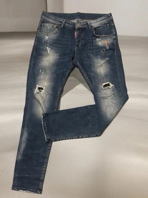 Dsquared2 Jeans  - Säljer ett par blå jeans med slitningar och hål på båda benen. Jeansen har en tvättad look med ljusa partier och små färgstänk samt en orange siffra 2 broderad vid fickan. 