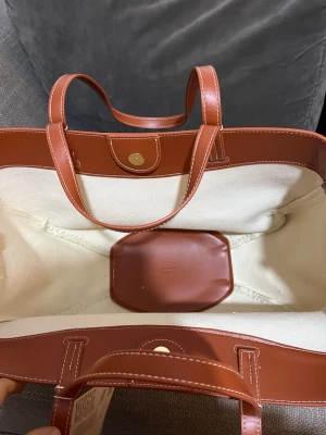 Shoppingväska i canvas och brunt läder - Snygg shoppingväskaka i beige canvas med detaljer och handtag i brunt läder. Väskan har en rundad form, vita sömmar och en liten innerficka. Perfekt för dig som vill ha en stilren och rymlig väska till vardagen.