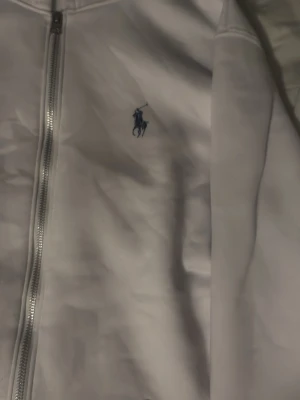 Vit hoodie från Polo Ralph Lauren - Vit hoodie med dragkedja från Polo Ralph Lauren. Klassisk design med blå broderad logga på bröstet, huva och stora fickor framtill. Perfekt för en clean och stilren look. Mjuk bomullskänsla och ribbade muddar vid ärmslut och nederkant.