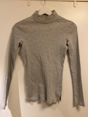Grå ribbad polotröja - Slimmad grå polotröja i ribbat material med långa ärmar och hög krage. Perfekt att styla lager på lager eller bära som den är.