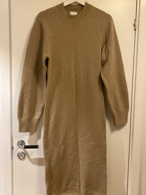 Beige stickad långklänning - Mysig beige stickad långklänning med rund halsringning och långa ärmar. Klänningen har ribbade muddar vid ärmslut och hals, samt en rak och enkel siluett.