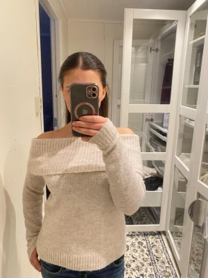 Beige off shoulder stickad tröja - Trendig beige stickad tröja med off shoulder-design och bred krage. Mjuk och skön, perfekt för lager-på-lager-look. Långa ärmar och ribbade muddar ger en snygg passform. Passar till jeans eller kjol för en chill stil.