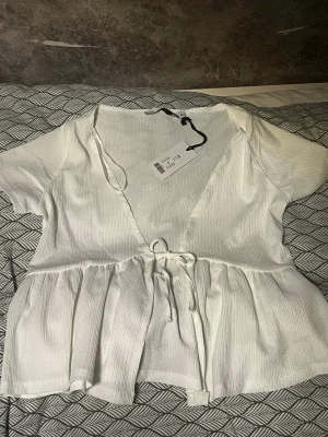 Vit omlottblus med knyt i midjan XS - Supersöt vit omlottblus i storlek XS. Blusen har korta ärmar, v-ringad hals och knytband i midjan som ger en snygg siluett. Materialet är lätt och har en diskret ribbad struktur. Perfekt för dig som gillar en feminin och fräsch look.