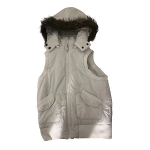 Vit väst med fuskpäls och huva - Snygg vit väst med avtagbar huva och fuskpälsdetalj. Västen har två stora fickor med lock och knappar, ribbad nederkant och dragkedja framtill. Perfekt för lager-på-lager och har en cool, sportig vibe. Storlek M men sitter mer som S