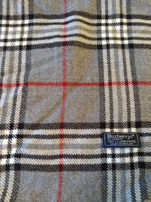 Rutig grå halsduk från Burberrys - Klassisk halsduk från Burberrys i 100% lammull med ikoniskt rutigt mönster i grått, svart, vitt och röda linjer. Mjukt material och snygga fransar i ändarna. Perfekt för att ge din outfit en tidlös och cool touch.