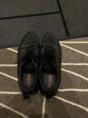 Svarta sneakers från Hugo Boss - Säljer ett par svarta Hugo Boss sneakers med diskret mönster på sidan och logga i rött på snörningen.
