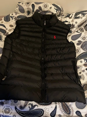 Svart dunväst från Polo Ralph Lauren - Svart dunväst från Polo Ralph Lauren med röd broderad logga på bröstet. Västen har quiltad design, hög krage och dragkedja framtill samt två sidofickor med dragkedja. Perfekt för lager-på-lager och snygg till streetwear.