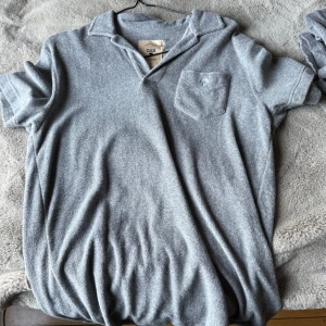 Grå frottéskjorta från OAS, stl S - Gråmelerad kortärmad skjorta från OAS i mjuk frotté. Skjortan har klassisk krage, bröstficka med broderad palm och avslappnad passform. Perfekt för chill dagar och sommarhäng.