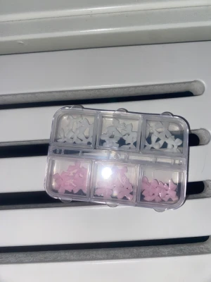 Söta rosetter för nageldekoration - Små dekorativa rosetter i ljusrosa och vit plast, perfekta för att pimpa naglarna. Kommer i en praktisk genomskinlig förvaringsbox med sex fack. Rosetterna är tredimensionella och ger en gullig touch till din nagelstil.