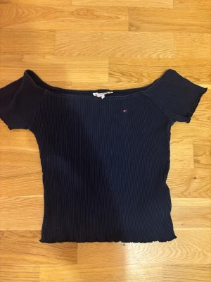 Mörkblå offshoulder topp Tommy Hilfiger - Snygg mörkblå ribbad topp från Tommy Hilfiger med offshoulder-design och korta ärmar. Toppen har en liten broderad logga på bröstet och vågiga kanter vid ärmslut och nederkant. Perfekt för dig som gillar stilrena och trendiga plagg.