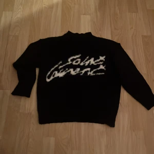 Svart stickad Saint Laurent tröja - Svart stickad tröja från Saint Laurent med vit, handskriven logga framtill. Tröjan har långärmad passform och en något högre hals. Perfekt för dig som gillar statement-plagg och exklusiva märken.