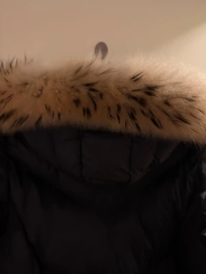 Svart Moncler dunjacka med päls - Säljer en lång, svart Moncler dunjacka med fluffig beige päls runt huvan och bälte i midjan. Jackan har Moncler-logga på ärmen, dragkedja framtill och quiltad design. Perfekt för kalla vinterdagar och riktigt snygg siluett.