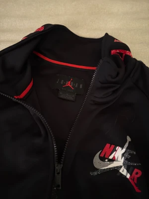 Svart Nike Air Jordan zipjacka - Säljer en svart zipjacka från Nike Air Jordan med röda och vita detaljer. Jackan har dragkedja, fickor framtill och texten 'AIR JORDAN' längs ärmarna. På bröstet finns ett stort Nike Air-tryck med Jumpman-loggan. Perfekt för dig som gillar sportig streetstyle.