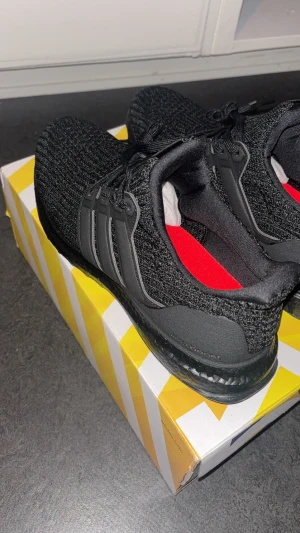 Svarta Adidas Ultraboost sneakers - Säljer ett helt nytt par svarta Adidas Ultraboost sneakers med stickad ovandel och klassiska tre ränder på sidan. Skorna har svart sula, snörning och röd innersula med Primeblue-tryck. Perfekta för dig som gillar stilrena och bekväma sneakers i syntetmaterial.