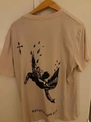 Beige t-shirt med svart tryck - Säljer en beige t-shirt med svart grafiskt tryck på ryggen och ett kors på bröstet. T-shirten har rund hals och korta ärmar. Materialet känns mjukt och skönt, troligen bomull. Texten 'REVELATION 12:9' finns längst ner på ryggen.