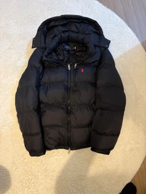 Svart pufferjacka från Polo Ralph Lauren - Svart pufferjacka från Polo Ralph Lauren med huva och tvåvägsdragkedja. Jackan har ett rött Polo-logo broderat på bröstet och är vadderad för extra värme. Perfekt för kalla dagar och har en clean, stilren look. Använd 2 gånger 