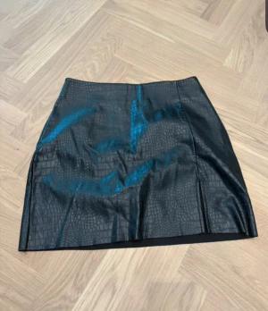 Svart kort kjol i skinnlook från Gina Tricot - Snygg svart kort kjol från Gina Tricot i skinnimitation med croco-mönster. Kjolen har en slits framtill och dragkedja i sidan för enkel påklädning. Perfekt för dig som gillar edgy och trendiga plagg med cool textur.