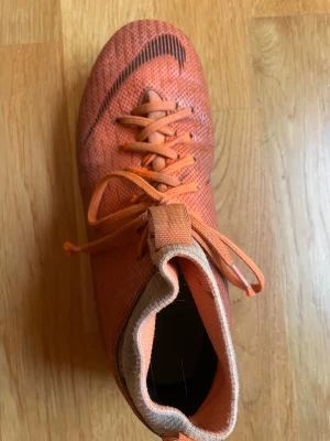 Nike Mercurial orange fotbollsskor - Säljer ett par Nike Mercurial fotbollsskor i orange med svarta detaljer och snörning. Skorna har en rund tå och platt sula med dobbar för gräsplan. Ovandelen är tillverkad i syntetmaterial med textur för bättre bollkontroll. Perfekt för dig som vill sticka ut på planen.