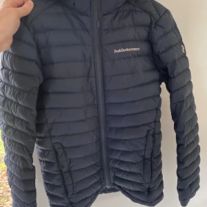 Mörkblå dunjacka från Peak Performance - Säljer en mörkblå dunjacka från Peak Performance, Modell JR FROST DH. Jackan är tillverkad i slitstark syntet med dunfyllning och har en diskret logga på bröstet och på ärmen. Den har en lite lagning på ena skuldran och en söm i quiltningen har släppt på ett ställe på ryggen (se bilder). Annars i mkt bra skick. Defekterna speglar det låga priset. Storlek 160. Modell JR FROST DH. 