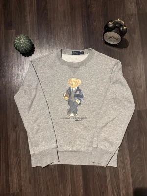 Grå sweatshirt Polo Ralph Lauren - Grå sweatshirt från Polo Ralph Lauren med klassisk Polo Bear-tryck på bröstet. Tröjan har rund hals, ribbade muddar och är långärmad. Mjuk insida och avslappnad passform, perfekt för chill dagar. Ikonisk och stilren design som alltid funkar. Nypris 3000