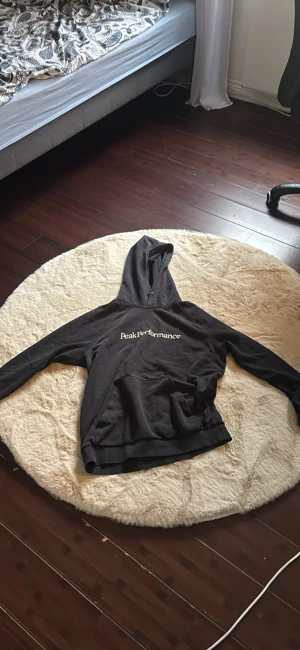 Svart hoodie från Peak Performance - Svart hoodie från Peak Performance med vit logotyp på bröstet. Tröjan har huva, lång ärm och en stor magficka framtill. Mjuk och skön, perfekt för chill eller träning. Klassisk loose passform och stilren design som funkar året runt.