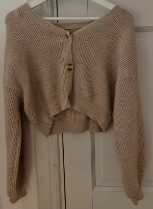 Beige/Brun croppad kofta med knappar - Säljer en beige/brun croppad kofta med ribbad stickning och snygga guldfärgade knappar framtill. Koftan har långärmad men kortare design och en avslappnad loose passform som ger en chill vibe. Perfekt för lager-på-lager under höst och vår.